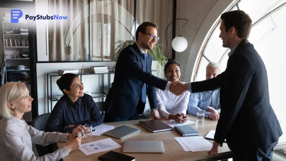 An employer shakes hands with a new employee in an office surrounded by coworkers and paperwork