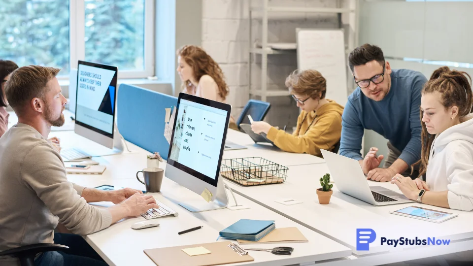 Employees in an office sit at desks, working on computers as part of a busy organization