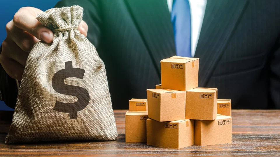 A person in a suit holds a burlap money bag next to stacked mini cardboard boxes.