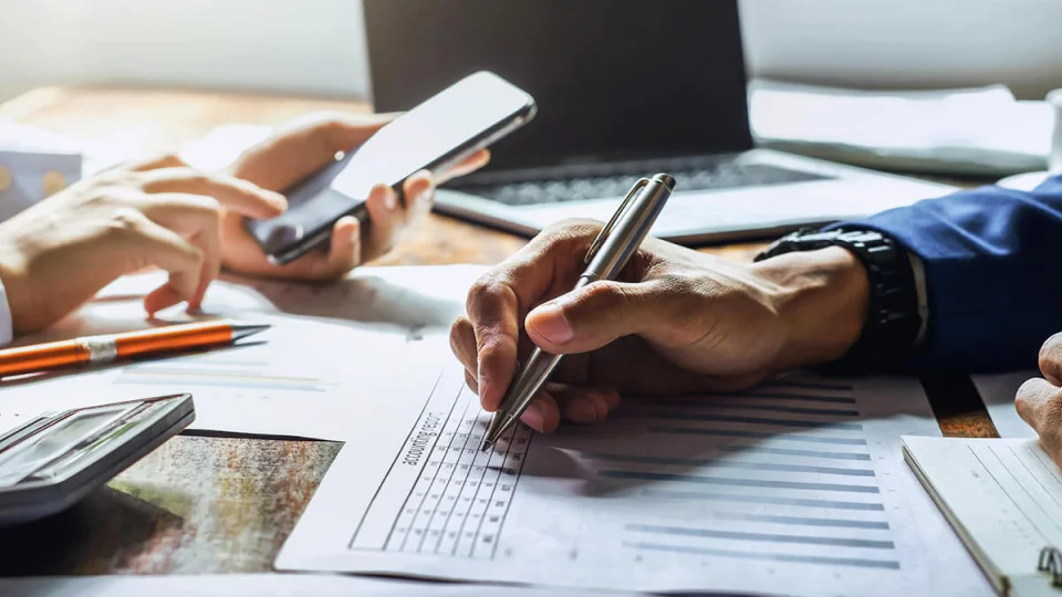 Two people sit at a desk reviewing an expense document together with a laptop and folders in the background.