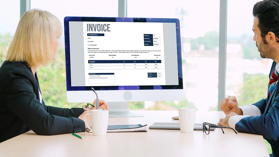Two people sit at a desk reviewing an invoice displayed on a large desktop monitor.
