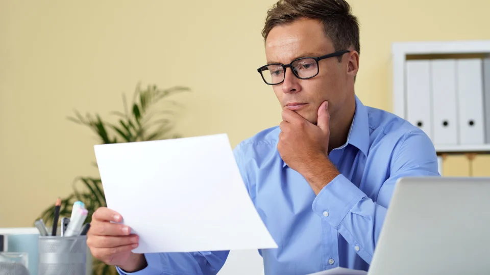 A man in glasses looks at an invoice while seated at a desk with a laptop.