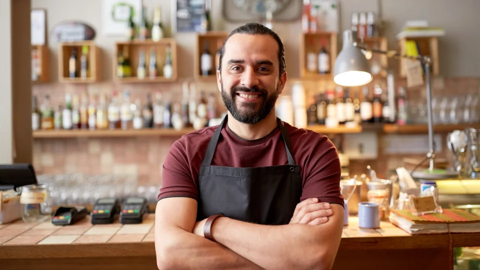 A small business owner stands behind a counter with arms crossed and smiling.