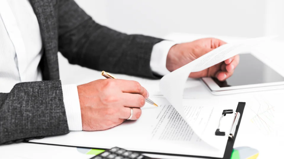 A person in a suit reviews and signs business documents on a clipboard.