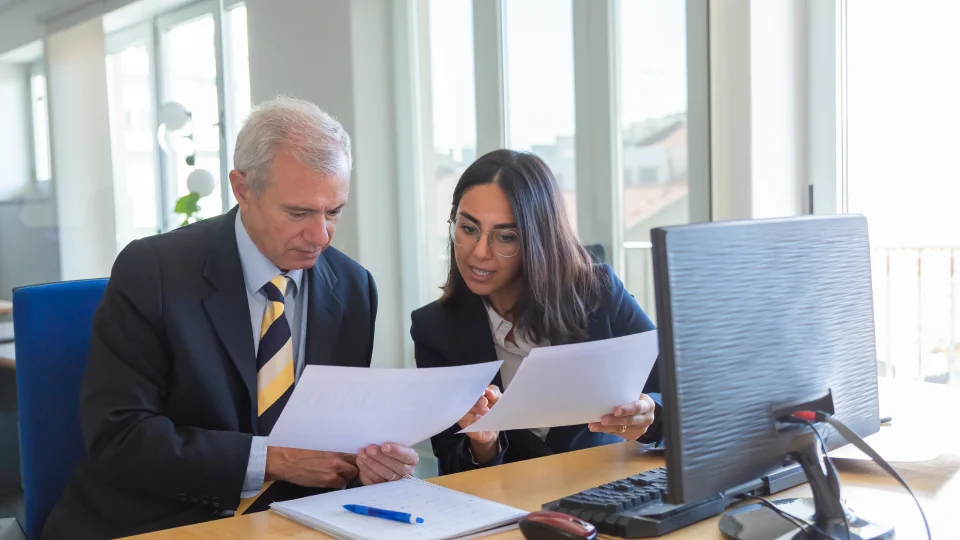 Two professionals sit at a desk reviewing financial documents together next to a computer.