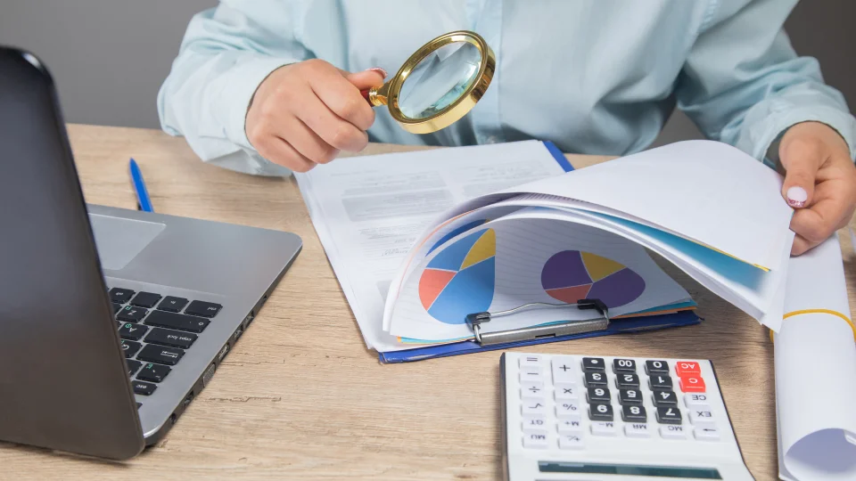 A person uses a magnifying glass to review financial charts at a desk.