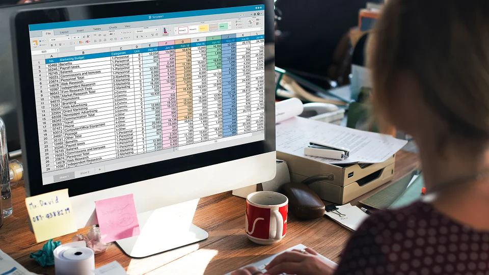 A person works at a desk with a computer displaying a color-coded spreadsheet.