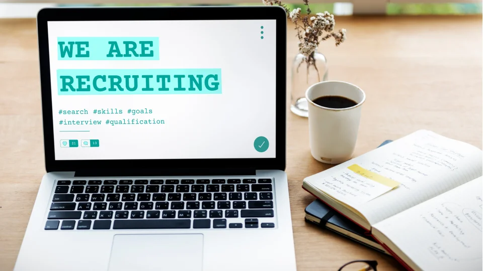 A laptop screen displays a recruiting message on a desk with a notebook and coffee.