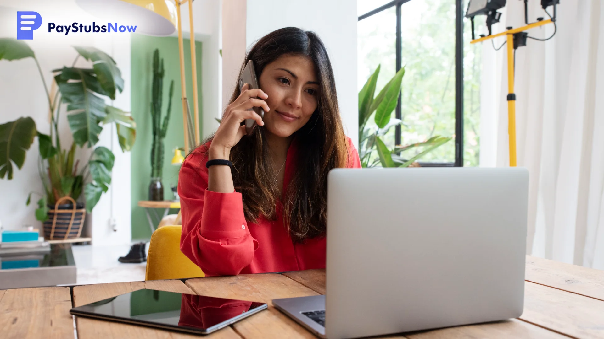 A woman in a red shirt is seated at a wooden desk with a laptop and tablet, smiling while talking on a cell phone in a bright room.